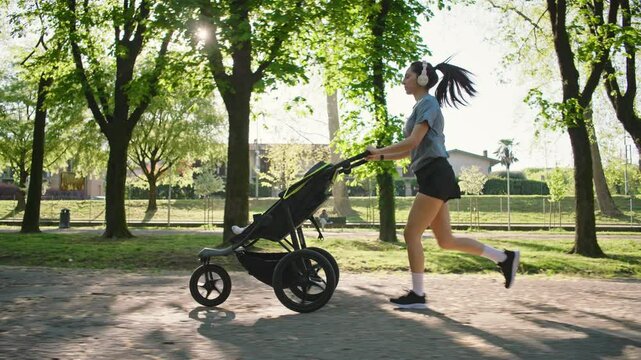 A young sporty woman runs in the park with her son in the running board. Mom is happy to do sports in the park with her newborn baby on a sunny day. Concept of power, fitness, family