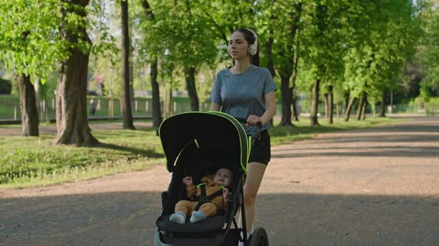 A young sporty woman walks in the park with her son in the running board. Mom is happy to do sports in the park with her newborn baby on a sunny day. Concept of power, fitness, family