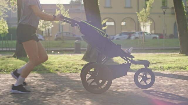 A young sporty woman runs in the park with her son in the running board. Mom is happy to do sports in the park with her newborn baby on a sunny day. Concept of power, fitness, family