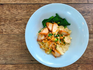 Bowl of noodles on wooden table. Noodles with roast pork and boiled dumplings served in blue bowl.  Noodles without soup garnished with sliced ​​roast pork, shrimp dumplings, Chinese kale,fried garlic