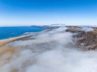 Obraz premium Aerial view of Rauðasandur, Iceland, where golden sands meet turquoise waters, framed by dramatic cliffs and low-hanging clouds under a radiant blue sky.