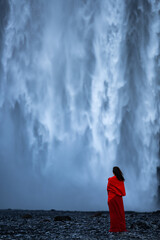 A woman in a red dress stands before the powerful Skógafoss waterfall in Iceland, her figure contrasting sharply against the mist and roaring cascade.
