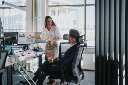 A multicultural team of business employees in an office meeting. A woman presents data on a screen to a seated colleague. The bright modern office fosters collaboration and productivity.