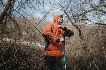Person wearing an orange jacket navigates through a dense forest, utilizing a trekking pole for...