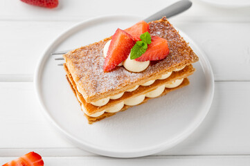 French dessert millefeuille with vanilla cream and fresh strawberries on a white plate on a white wooden background.