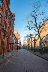 A red brick paved street in Manchester, England, lined with red brick buildings, bare trees, and a...