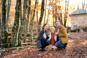 Smiling dad and mom are squatting near a little girl among fallen leaves in the forest