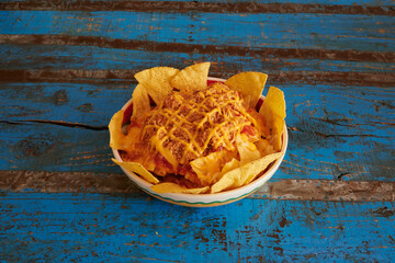 A bowl of nachos with cheese and salsa. The bowl is on a blue table. The nachos are piled high and covered in cheese