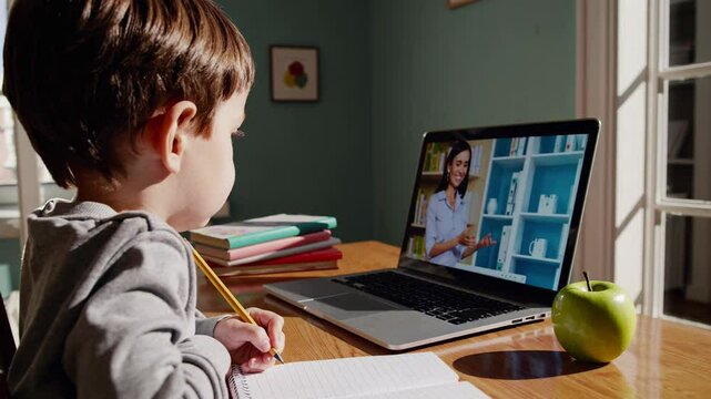 Preschool boy writing in notebook during online class, sitting at desk with laptop, teacher visible on screen and apple nearby. Home training