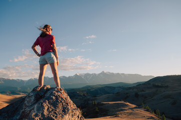 A woman in casual clothing stands on a rocky peak, gazing at scenic mountains and a vast valley...