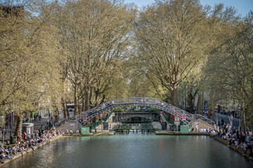 Paris, France - 04 05 2025: Canal Saint-Martin. Panoramic view of people sited, apartment buildings, a bridge and trees reflecting on the water.