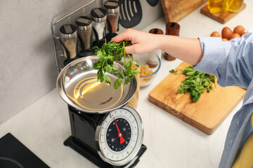 Woman putting parsley into bowl on mechanical kitchen scale at white marble countertop indoors,...