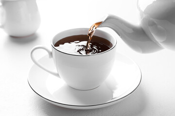 Pouring freshly brewed tea from teapot into cup at white table, closeup