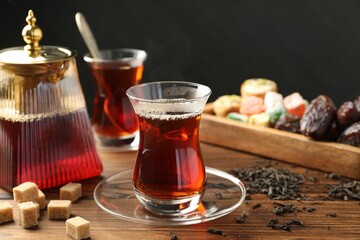 Traditional Turkish tea in glass cups, teapot, dry leaves, brown sugar and sweets on wooden table, closeup