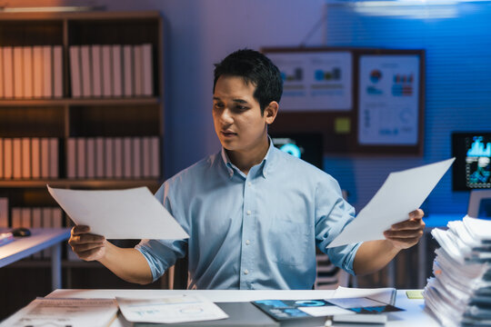 Young professional comparing two documents while sitting at his desk in a dimly lit office, working late at night