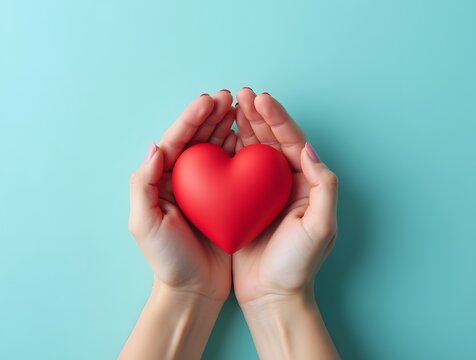 First person top view photo of female hands cradling a red heart on a pastel blue background. There's empty space for text or an ad. It's a concept for World Blood Donor Day - Powered by Adobe