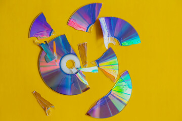 Pieces of a shattered and broken compact disc are scattered in different directions on a yellow background, viewed from above. A cracked CD disc, damage to an old storage medium, outdated technology.