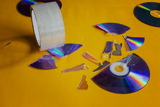 Pieces of a broken compact disc on a light yellow background with tape trying to fix the break, close-up shot, damage to an old storage medium, vintage technology from past years, archive restoration.