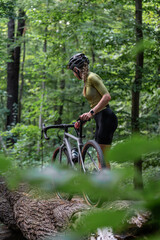 Female cyclist in full cycling gear, including helmet, stands with her gravel bike in midst of dense forest. Rear view. concept of outdoor exploration, healthy living.