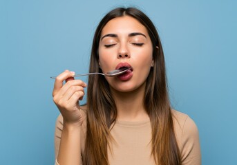 Obraz premium A woman with eyes closed, enjoying the taste of something from a spoon against a blue backdrop.