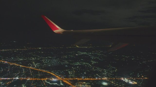 Aerial view of urban city skyline at night with glowing lights, captured from airplane window showing wing and illuminated streets, creating mesmerizing travel scenery.