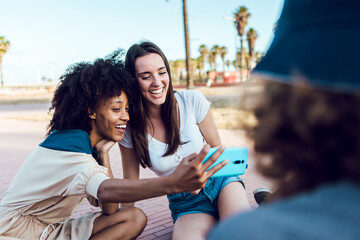 women friends taking a selfie with smart phone outdoors - Joyful multiethnic female couple having fun with their friends in summer holidays. Millennial generation, friendship and youth concept