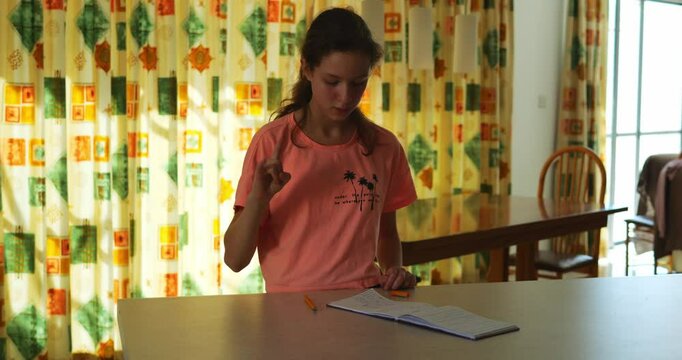 Thoughtful young weoman carefully repeats sequence of sign language hand gestures. Young brunette closely examines notes standing near table against window curtain