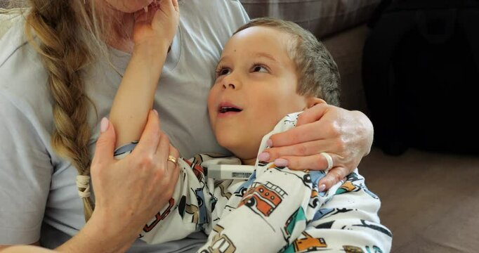 Sick little son smiles and plays with mom tongue while measuring temperature. Woman cheers up preschooler boy with fever hugging and holding hand