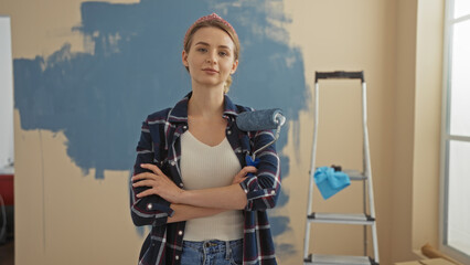 Woman painting new home stands confidently with crossed arms in living room holding roller brush with ladder nearby, surrounded by fresh blue wall paint.