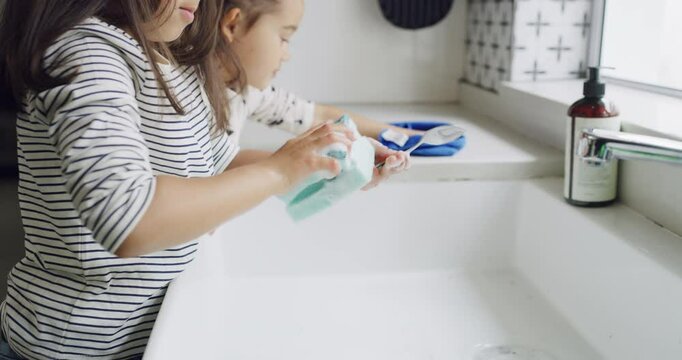 Sisters, kids and washing dishes in kitchen, cleaning or learning hygiene in home. Children, soap and spoon at sink for housework, chores and girls together to help wipe counter with cloth and sponge