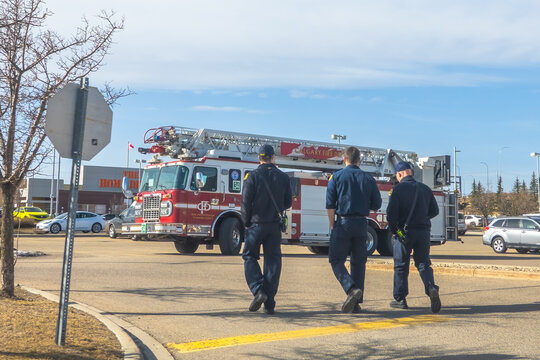 Calgary, Alberta, Canada. Apr 13, 2025. Three firefighters in uniform walk towards a Calgary Fire Department truck with an extended ladder in a parking lot under a partly cloudy sky.