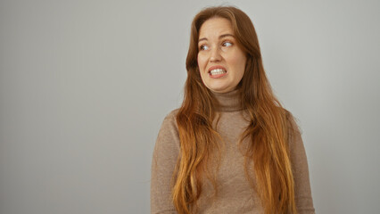Young woman with red hair makes a face while standing in front of an isolated white background wall showcasing an uncertain expression in a casual setting.