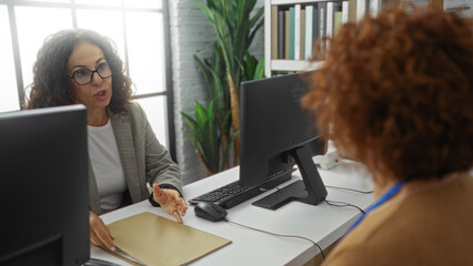 Obraz premium Two hispanic businesswomen in an office discuss work over a desk with computers and documents, indicating collaboration and professional partnership indoors.