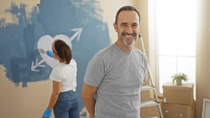 Middle-aged hispanic couple painting heart on new home wall, man smiling while woman paints, symbolizing love, family, and fresh beginnings in cozy indoor setting.