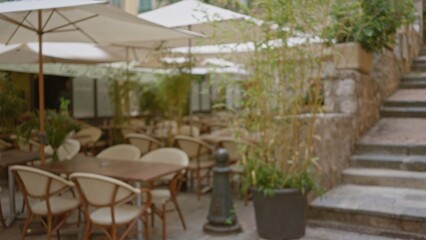Blurred outdoor cafe with empty chairs and tables under umbrellas in villefranche-sur-mer, france, surrounded by greenery and rustic stone walls.