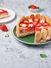 Round tiramisu cake with fresh strawberries and mascarpone cream decorated with marshmello flowers on a green plate on a light marble background. No-bake desserts.