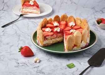 Round tiramisu cake with fresh strawberries and mascarpone cream decorated with marshmello flowers on a green plate on a light marble background. No-bake desserts.
