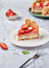 Sliced ​​round tiramisu cake with fresh strawberries and mascarpone cream decorated with marshmello flowers on a green plate on a light marble background. No-bake desserts. Italian cuisine