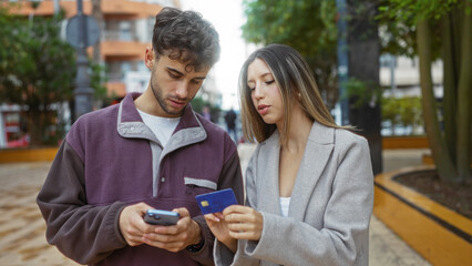 Man and woman in casual attire using phone and credit card discussing plans in an urban outdoor setting with modern architecture visible in the background.