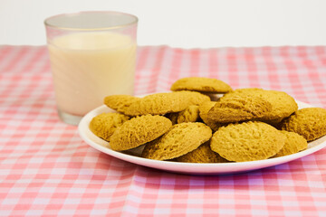 A plate of cookies sits on a pink and white checkered tablecloth next to a glass of milk. The cookies are arranged in a pyramid shape, with the top cookie being the largest
