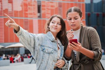 Two young women engaged in an animated conversation while one points excitedly, sharing insights and directions with a smartphone in hand, reflecting a moment of genuine interaction and curiosity