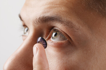 Man putting color contact lens in his eye on light background, closeup
