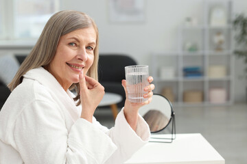 Smiling woman with glass of water taking collagen pill at home. Space for text