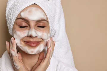Beautiful woman washing her face with cleansing foam on beige background, space for text
