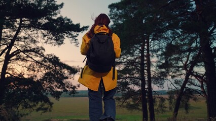 Woman with tourist backpack walks alone through forest to field after sunset. Woman traveller carries backpack on hike in autumn park. Sportive woman rests on solitude holiday to wild nature