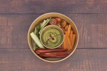 A bowl of vegetables and dip on a wooden table. The dip is green and the vegetables are carrots and cucumbers
