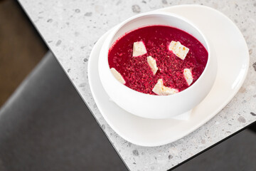 A vibrant bowl of beetroot soup topped with crumbled cheese and spices, served on a white plate on a speckled countertop, showcasing healthy and colorful cuisine.