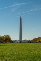 Fototapeta premium The Washington Monument rises prominently above the National Mall in Washington DC, framed by green grass, autumn trees, and a clear blue sky on a crisp fall day.