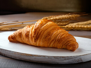 Golden-brown croissant on a wooden board with wheat stalks in the background, highlighting fresh...
