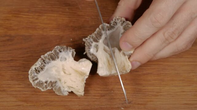 A cook cuts morel mushrooms into small pieces using a knife on a wooden cutting board. Close-up.